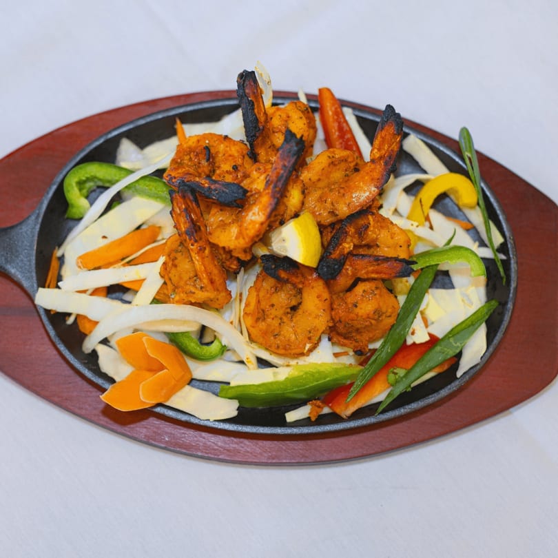 Sizzling tandoori shrimp with charred edges, lemon, and colorful vegetables on a cast iron platter.