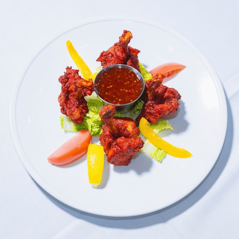 Red chicken lollipops with dipping sauce, lettuce, bell peppers, and tomatoes on a white plate.